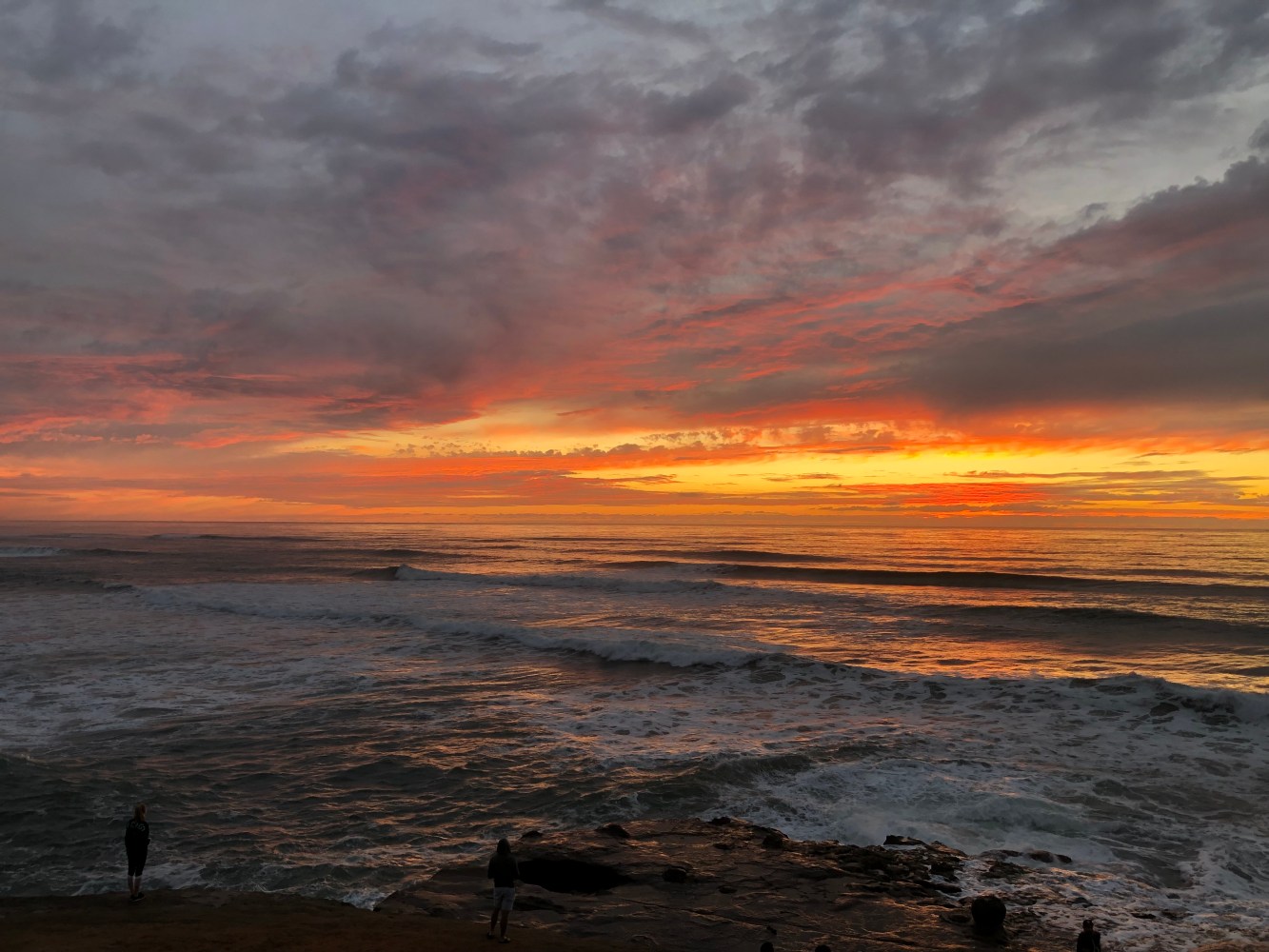 Sunset over ocean with clouds and waves, people on rocky shore.